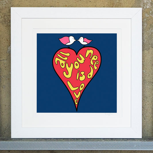 Giclee original artwork print in a white mounted frame. A bright red heart has the letters in yellow bubble writing of all you need is love. The background is navy blue, on top of the heart are two pink winged seagulls facing each other with their beaks touching, looking into each others eyes.