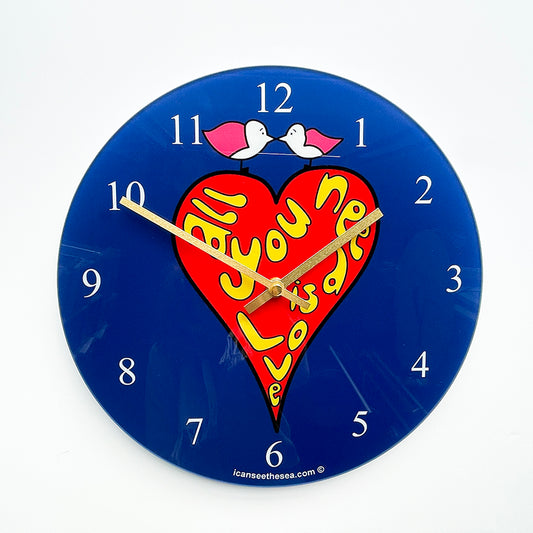 Recycled glass clock with yellow bubblewriting of all you need is love inside a red heart. On top of the heart are two pink winged seagulls facing each other. The background is blue