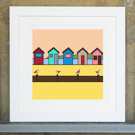 Giclee original artwork print in a white mounted frame. Five beach huts in different colours are on a bright yellow sandy beach. Towards the front are groynes with four pink winged seagulls perched on top looking in various directions. In the far background behind the beach huts is the blue sea with a few sailing boats in the distance with a green land and pale beach sky. 