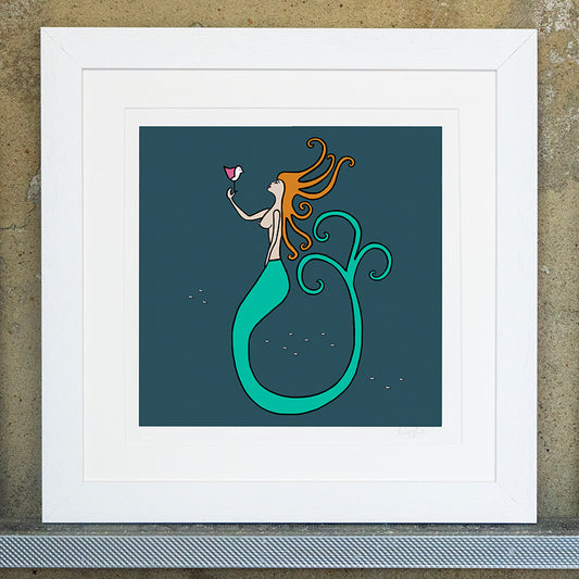 Giclee original artwork print in a white mounted frame. A mermaid with ginger hair and a long green tail is underwater surrounded by small little fishes. The background is teal and the mermaid is holding a pink winged seagull where they are looking at each other. The print is called mermaid kisses.