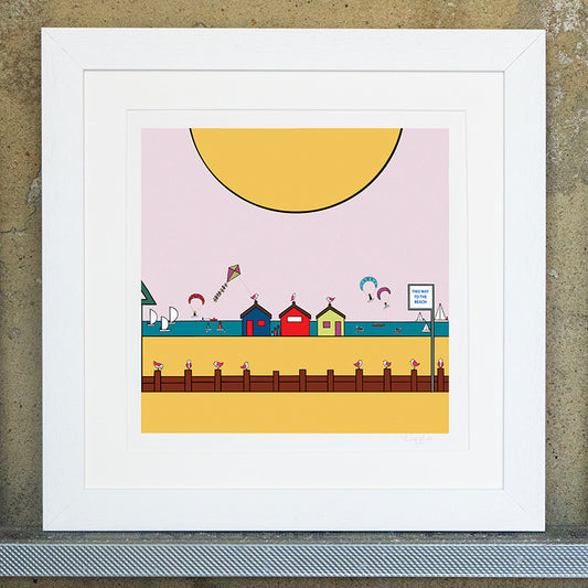 Giclee original artwork print in a white mounted frame. A pink sky and large yellow sun is at the top. Below is the beach and sea, with three small colourful beach huts with pink winged seagulls perched on top. The sea is busy with kites, small sailiing boats and water sports. At the front is golden sand with a sea defence with groynes and a sign that says this way to the beach. More small little pink winged birds are sat on top on the groynes. 