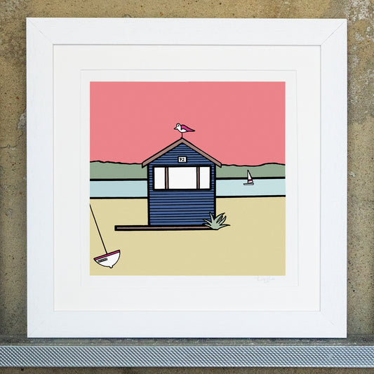 Giclee original artwork print in a white mounted frame. A navy blue beach hut with the number 72 has a pink winged seagull perched on top. The ground is a sandy colour with a small leaning boat. A small plant and wooden deck is at the bottom of the beach hut, with a landscape of the sea in the background, greenery and a warm pink sky. A small sailing boat is in the water. Each shape and detail is outlined in thick black.