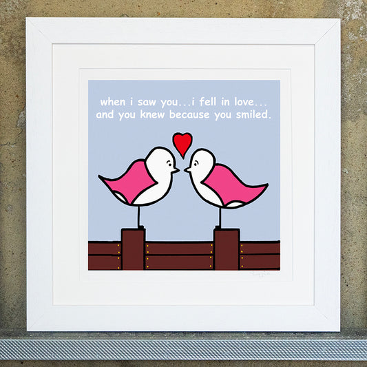 Giclee original artwork print in a white mounted frame. Two pink winged seagulls are facing each other stood on a groyne, above both their heads is a red love heart. The sky is pale blue with white text that says when i saw you...i fell in love...and you knew because you smiled.