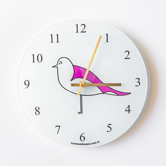 Recycled glass white face clock with a pink wing seagull in the centre brass coloured hands with black numbers and icanseethesea at the bottom