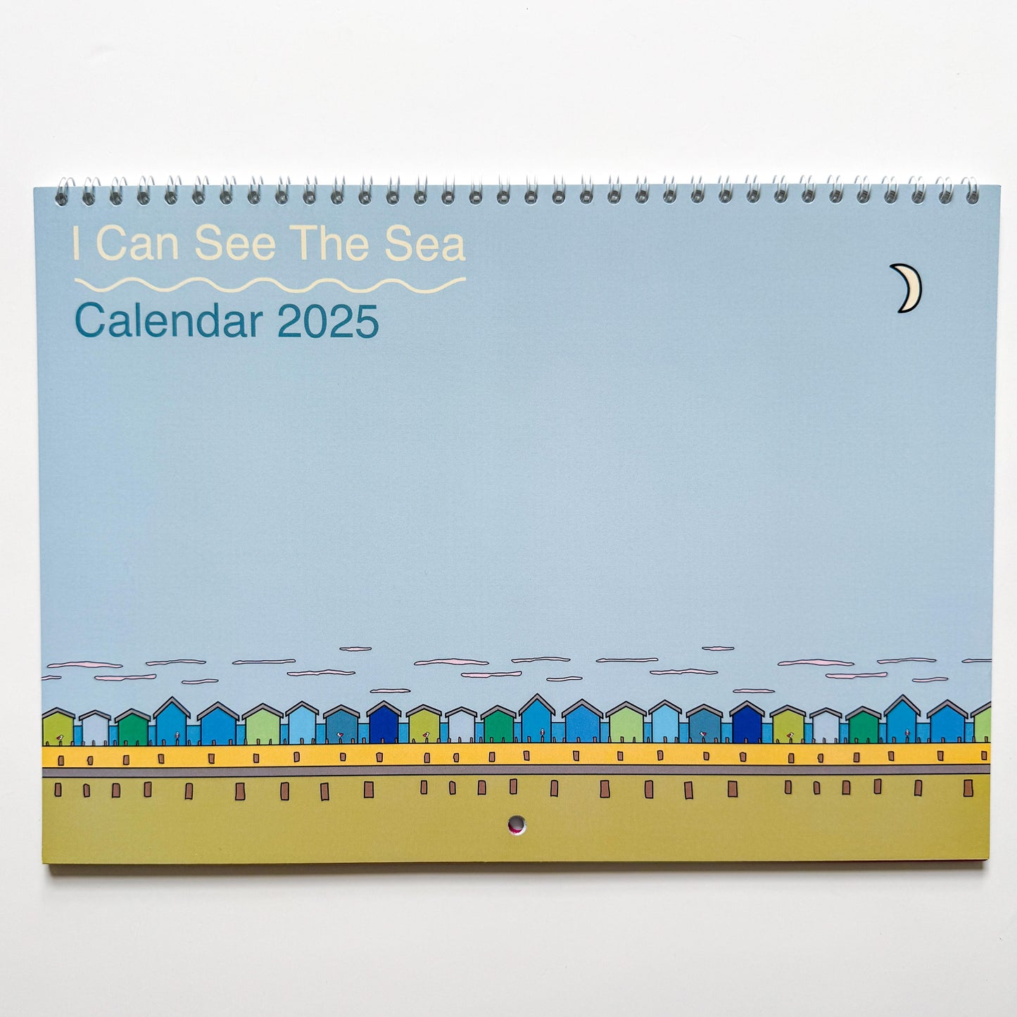 A ringbound calendar is on a white background. The calendar is light blue with blue and green beach huts on a sandy beach on the bottom. the top left corner says I Can See The Sea calendar 2025 in white and blue. In the right corner is a small white moon.