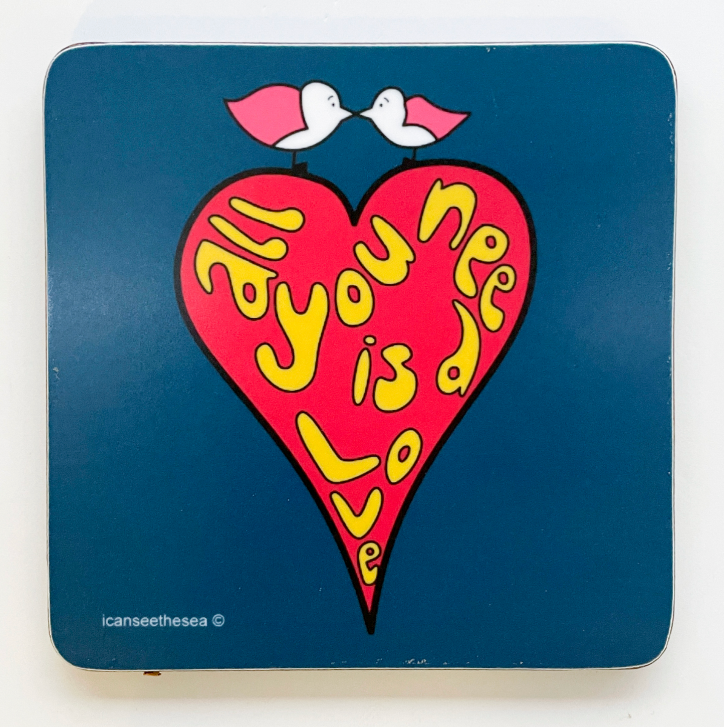 coaster with a navy blue background, in the centre is a red heart with all you need is love in yellow bubble writing. The heart is outlined in a black line. On top of the heart sits two pink winged seagulls facing each other and their beaks touching.