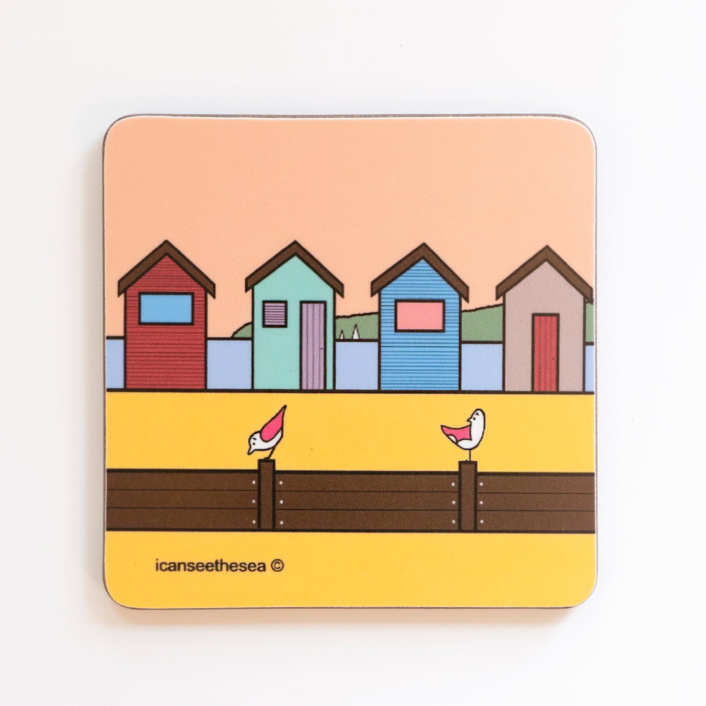 A coaster with bright warm colours. On the horizon is four different coloured beach huts, behind them is in the sea with two small sailing boats and a green landscape. The sky is a peach colour, in front of the huts is yellow sand with a brown fence, on top of the fence posts sits two pink winged seagulls looking in different directions.