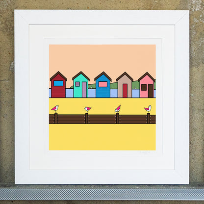 Giclee original artwork print in a white mounted frame. Five beach huts in different colours are on a bright yellow sandy beach. Towards the front are groynes with four pink winged seagulls perched on top looking in various directions. In the far background behind the beach huts is the blue sea with a few sailing boats in the distance with a green land and pale beach sky. 