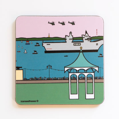 A coaster of southsea bandstand and the Queen Elizabeth air craft carrier. The colours are blues, greens and pinks. In the distance is the isle of wight in a green landscape, the sea is bright blue with small sailing and fishing boats. The sky is pink with three helicopters and in the sea the air craft carrier. On the sea's edge is a brick wall and little figures in different colours looking out. Southsea band stand sits at the front on a green field.