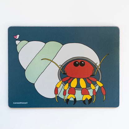 a placemat with a navy background of a painting of a hermit crab. The shell is stiped with pale green and pale blue. The crab is red and yellow with two black beady eyes. On the point of the shell is a pink winged seagull looking out. All the details and shapes are outlined in black.