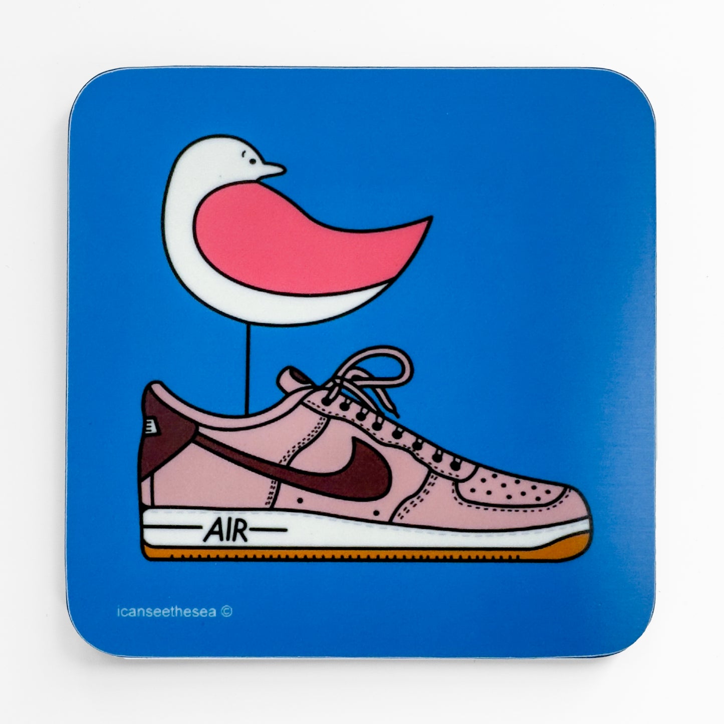 a coaster with a bright blue background has a print of an original painting of one air pink trainer. Inside the trainer stands a pink winged seagull looking back. All the shapes and details are outlined in black liner.