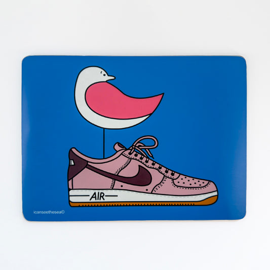 a placemat with a bright blue background has a print of an original painting of one air pink trainer. Inside the trainer stands a pink winged seagull looking back. All the shapes and details are outlined in black liner.