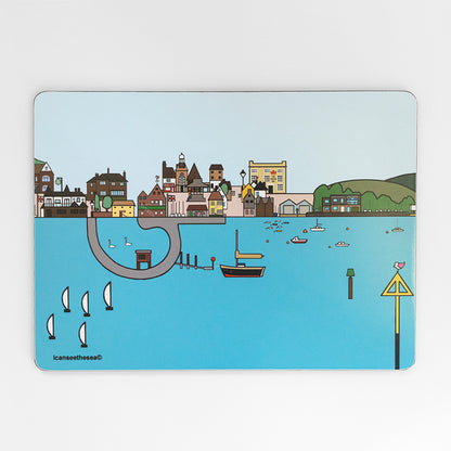 a placemat of a painting of emsworth. The water is pastel blue with sailing boats, swans, fishing boats and buoys. The horizon has lots of detailed buildings, streets, trees and a pub. The sky in the back ground is pale blue, and the water has a small area to walk round with swans. 