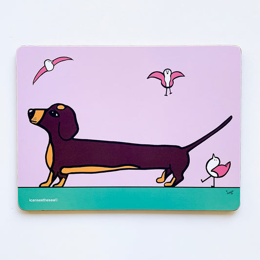 a placemat of a painting of a sausage dog called follow the leader. The sky is pink with a green ground, a brown sausage dog with beige patches has a pink winged seagull following behind. In the sky are two more pink winged seagulls flying. All the shapes and details are outlined in black liner.