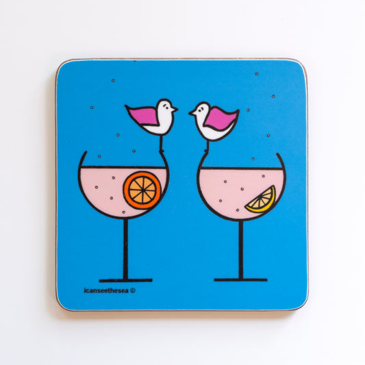 a coaster of a painting of two gin glasses. The liquid is pink and one has a lemon slice and the other an orange slice. The drink has small bubbles which float out of the glass and up. Two pink winged seagulls are sat on the rim of each glass looking at each other. The background is bright blue.