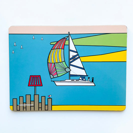 a placemat of a painting of a sailing boat with a colourful sail. The sea is bright blue, with yellow and green landscapes. In the far distance is three white sailing boats and a pale pink sky. At the bottom is yellow sand and a sea groyne with a pink winged seagull perched on top.