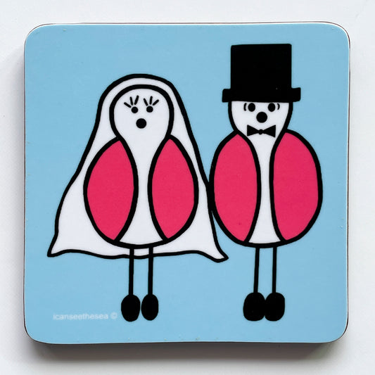 A pastel blue coaster with two pink winged seagulls stand side by side. One is wearing a grooms outfits of hat, bow tie and black shoes the other a wedding veil, big eyelashes and black shoes. The seagulls have white bodies with black eyes and round black nose.