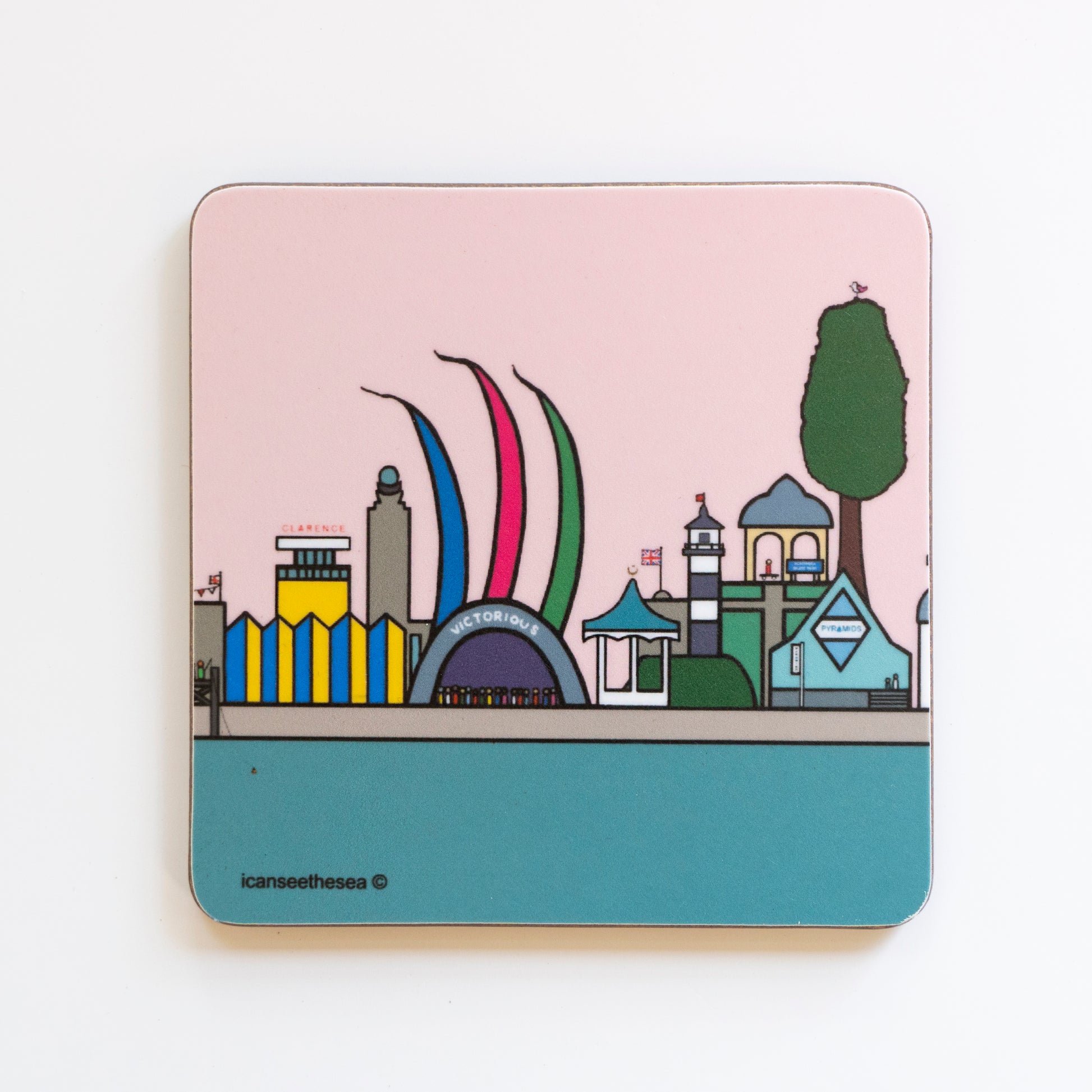 A coaster of a painting of victorious festival and southsea. The sea is teal with a horizon of different iconic buildings, clarence pier, southsea skate park, pyramids, ancient monument, southsea bandstand, southsea castle and the pier where a figure is fishing. The sky is pastel pink, and there are large colourful flags coming out of the victorious stage with a crowd of figures.