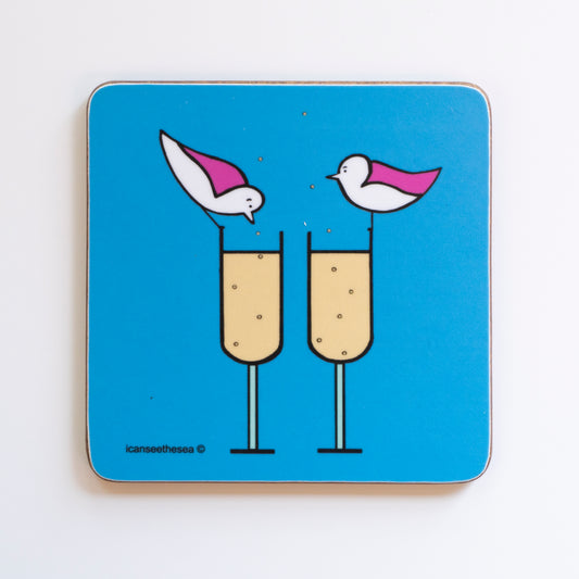 A bright blue coaster of a painting of champagne bubbles. Two champagne flutes are side by side with pale liquid and bubbles in the glass rising up out the top of the glass. Two pink winged seagulls are perched on each glass rim, one is looking down at the drink and the other looking across.