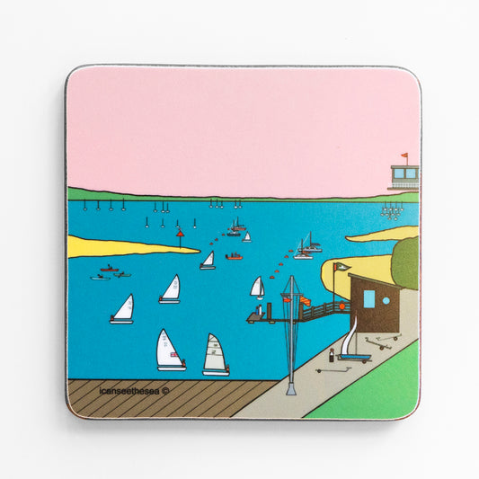 A coaster of Mengeham Rythe sailing club, with a bright blue sea with sailing boats, buoys and kayaks. There are yellow sandbanks, wooden decking, flag poles and small look out huts. In the horizon there is green representing the landscape and a pastel pink sky. Small figures can be found on the boats and waiting on the decks.