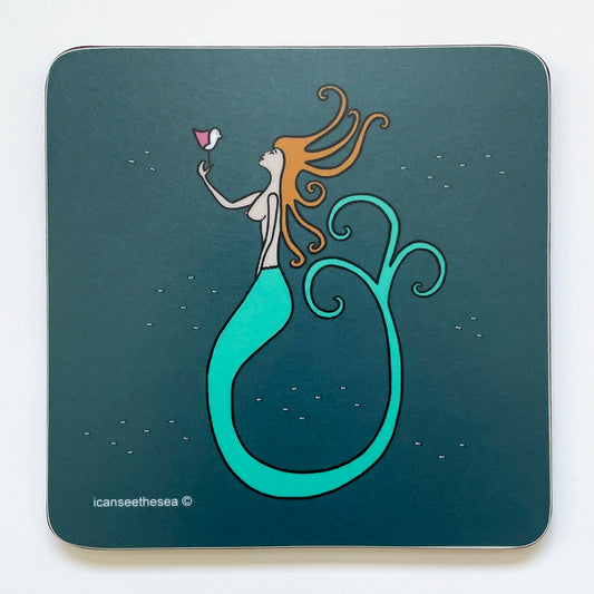 A coaster of a painting of a mermaid in the sea. The background is teal with small fishes swimming in small groups, a mermaid with golden floating hair that goes in different directions and some spiral. The mermaid is holding up a pink winged seagull where they look at each other. The mermaids tail is bright turquoise and curls upwards into three spiral curls. The mermaid is topless with pale skin, red lips and blue eyes.