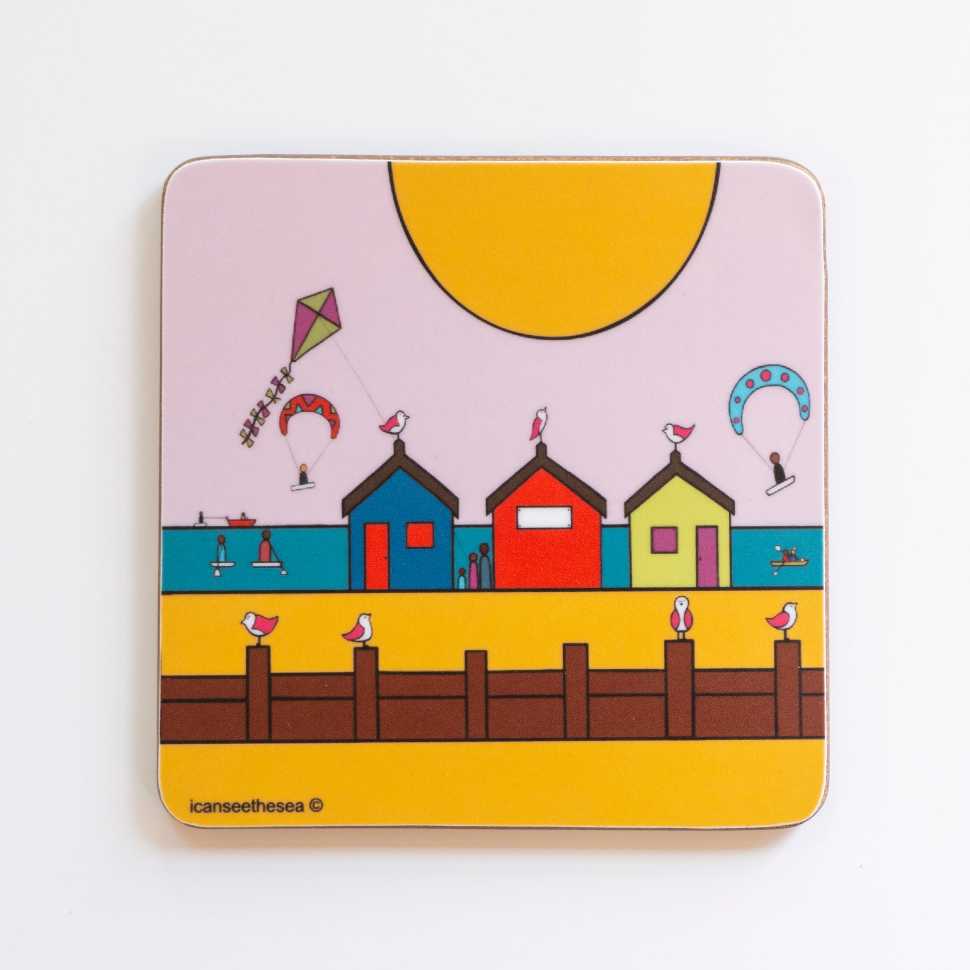 A coaster of a painting of Mill Rythe. The beach is golden yellow, with a wooden groyne where pink winged seagulls sit on top. In the distance are three brightly coloured beach huts with a pink winged seagull perched on top of each one. The sea is teal blue behind the huts where there is a kite flying, kayaks, canoeing and kite surfing with small figures. The sky is pale pink with a big golden yellow sun at the top.