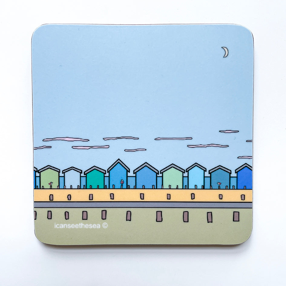 A pale blue coaster of a painting of the seaside. The sky is blue with a small little white half moon. At the bottom is the beach with wooden posts, a road and yellow sandy beach. In the distance is a row of different shades of blue and green beach huts. On the posts in front pink winged seagulls are perched. In the far distance is the blue sea and small white clouds in the sky.