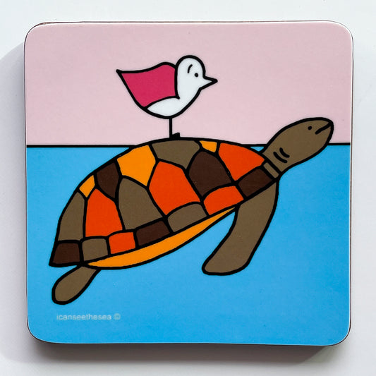 A coaster of a painting of a orange, red and brown turtle and pink winged seagull. The sea is bright blue with the turtle swimmming with its head above water, the seagull is perched on top of the turtles shell facing ahead. The sky is pastel pink and the turtle shell is colourful with pentagon shaped colours and its underbelly light orange.