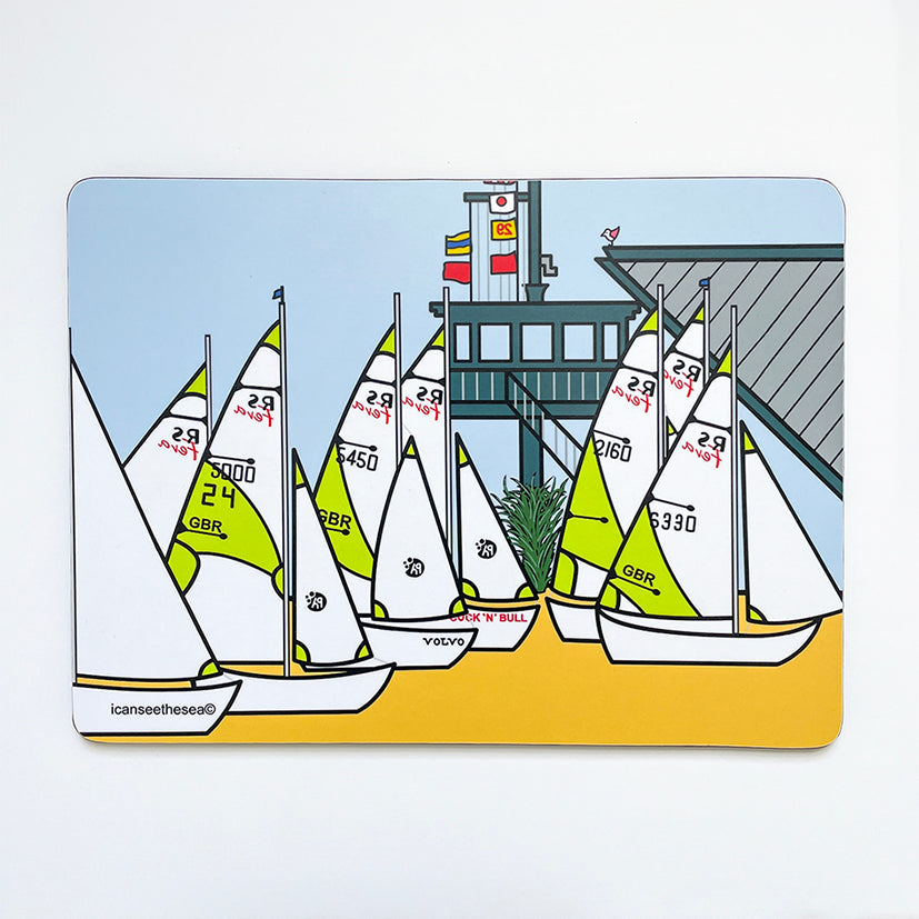 A placemat of sailing boats. The sand is bright yellow with sailing boats parked on the beach with lime green and white with numbers and logos. Behind them is a wooden lookout with steps, balcony and mast with flags. On top of the lookout sits a pink winged seagull. The sky is pastel blue and some green plants are growing round the bottom of the lookout under the steps.