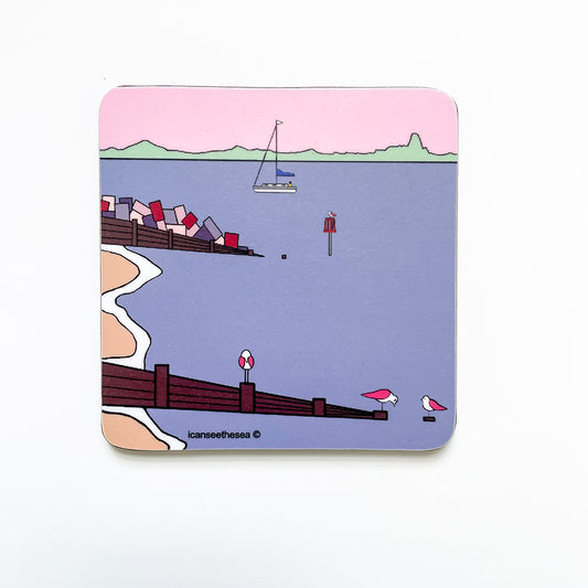 A coaster of a painting of sandy point. The sky is pastel pink and the sea mauve. The horzion has a green trim to represent trees, hedges and the landscape. In the water is a sailing boat and sea marker with a small pink winged seagull perched on top. Two sea groynes come in from the left with a sandy beach and white curves where the waves foam. There are three pink winged seagulls perched on the groynes at the front looking in different directions and positions.