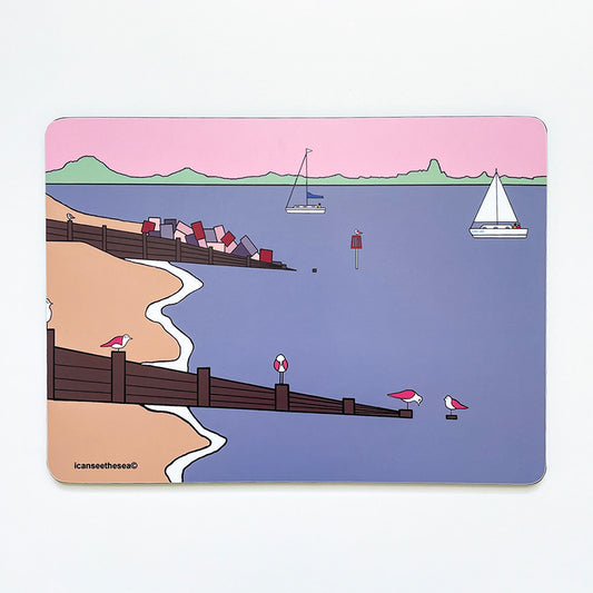 A placemat of a painting of sandy point. The sky is pastel pink and the sea mauve. The horzion has a green trim to represent trees, hedges and the landscape. In the water is a sailing boat and sea marker with a small pink winged seagull perched on top. Two sea groynes come in from the left with a sandy beach and white curves where the waves foam. There are three pink winged seagulls perched on the groynes at the front looking in different directions and positions.