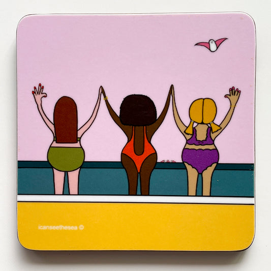 A coaster of a painting of three women on the beach. The sky is pale pink with a teal coloured ocean and bright yellow sandy beach. Three women in bikinis and swimsuit face the sea in a row with their arms up holdind hands. There is a pink winged seagull flying in the sky above.