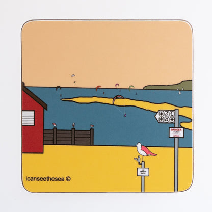 A coaster of a painting of a beach. The sky is a pale peach colour, with blue sea, yellow sand and green landscape on the horizon. To the left is a red beach hut with brown sea groyne. In the distance are figures windsurfing out at sea with sailing boats. On the beach is a tap with a pink winged seagull sat on top and a pole with a sign.