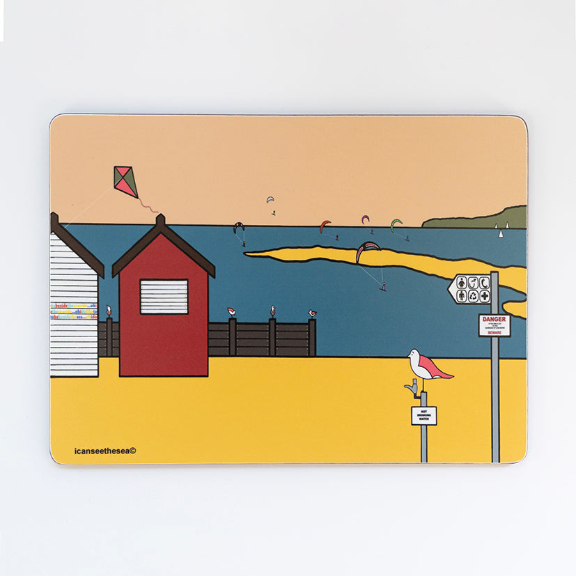 A placemat of a painting of a beach. The sky is a pale peach colour, with blue sea, yellow sand and green landscape on the horizon. To the left is a red beach hut with brown sea groyne. In the distance are figures windsurfing out at sea with sailing boats. On the beach is a tap with a pink winged seagull sat on top and a pole with a sign.