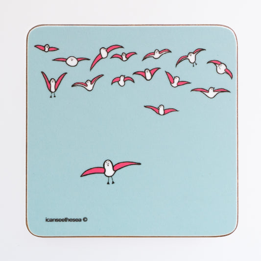 A coaster of a painting of seagulls windsurfing in the sky. The background is a pale blue green colour with half a dozen pink winged seagulls flying in the sky. One pink winged seagull is slightly lower than the rest in flight. All the seagulls are looking different directions.