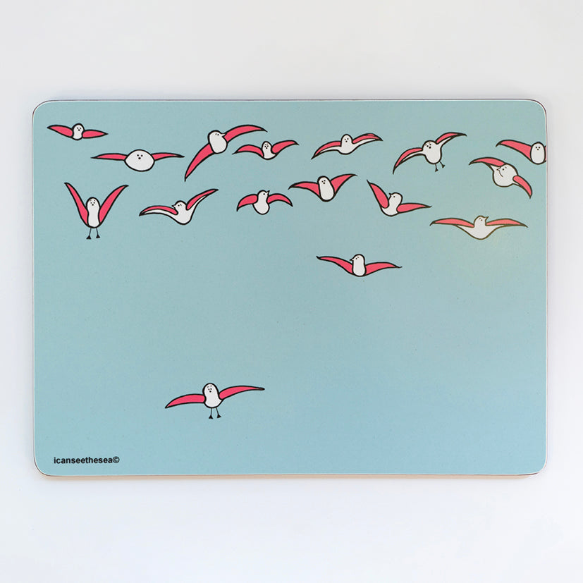 A placemat of a painting of seagulls windsurfing in the sky. The background is a pale blue green colour with half a dozen pink winged seagulls flying in the sky. One pink winged seagull is slightly lower than the rest in flight. All the seagulls are looking different directions.