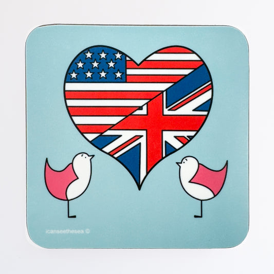 A coaster of a painting of a big heart with two pink seagulls. The background is a pale blue colour with a large heart in the centre filled with a british and american flag. Either side of the heart are two pink winged seagulls looking up at the heart.