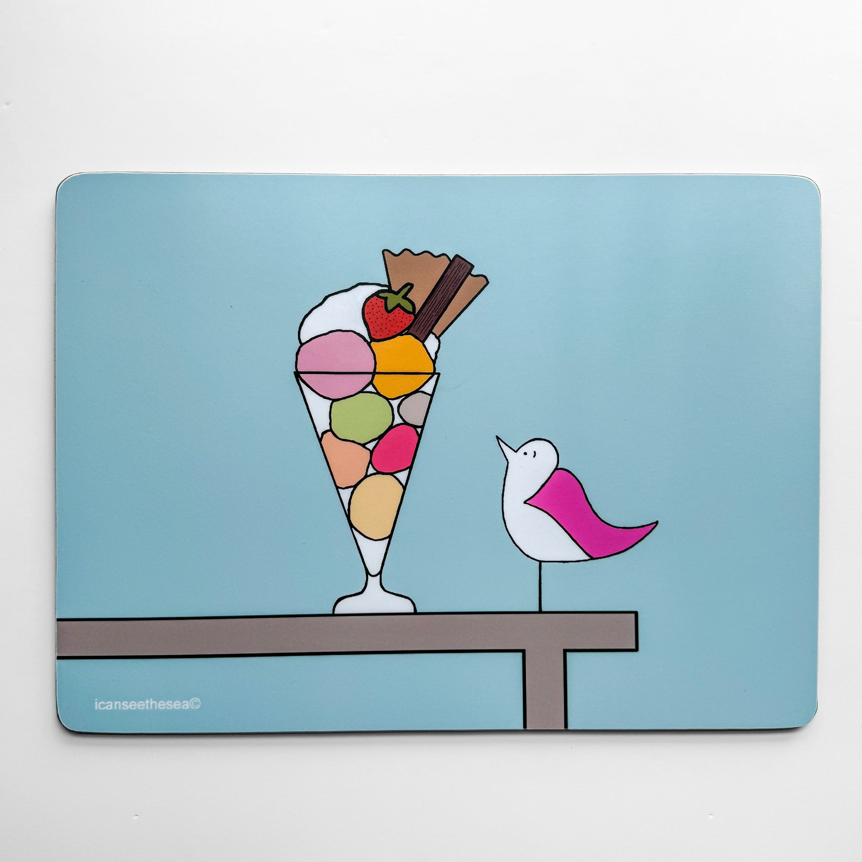 Placemat with light blue background, with a design in the centre of a wooden table with an icream sundae with strawberries, cream, flake and different coloured icream scoops. On the table a pink winged seagull sits looking up at the dessert which is in a tall glass. The shapes and details are outlined in a fine black line.